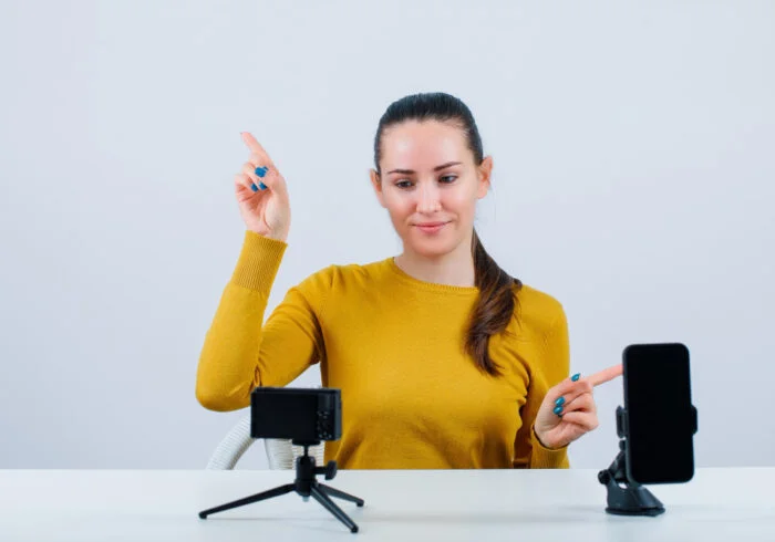 Como criar vídeos profissionais: Dicas para iniciantes 4 smiling blogger girl is pointing up right with forefingers by sitting front her mini camera white background