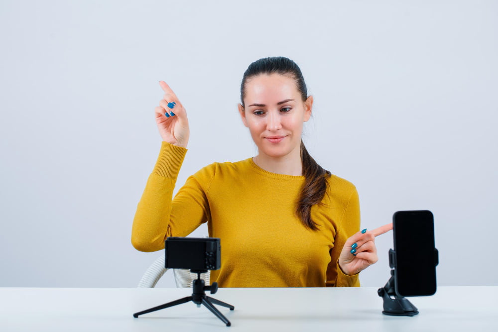 smiling blogger girl is pointing up right with forefingers by sitting front her mini camera white background